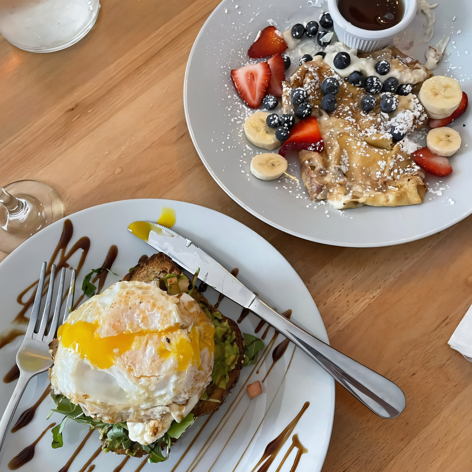 Avocado toast with a fried egg and fruit crepes with syrup on a wooden table.
