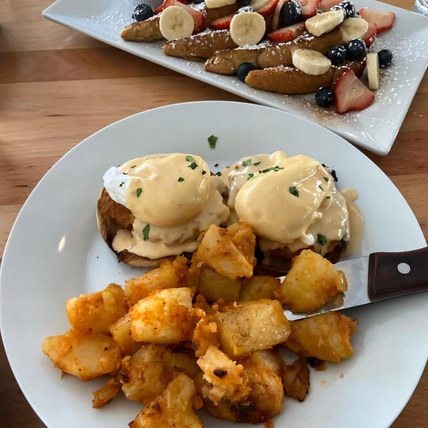 Eggs Benedict with home fries and French toast with fruit and powdered sugar on a wooden table.
