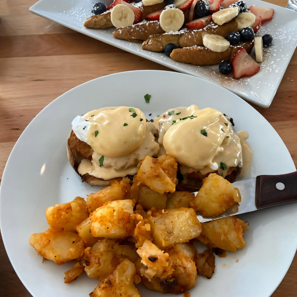 Eggs Benedict with home fries and French toast with fruit and powdered sugar on a wooden table.