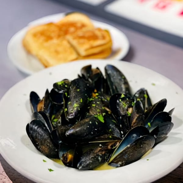 A white bowl of steamed mussels with parsley and broth, with toasted bread in the background.