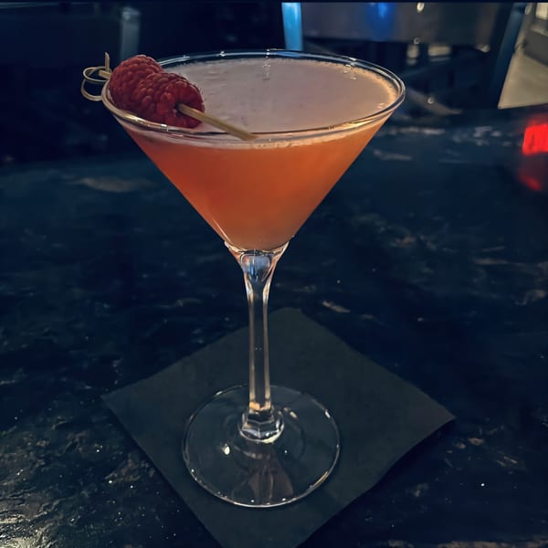 A vibrant orange cocktail in a martini glass, garnished with raspberries on a dark bar.